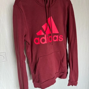 Vinröd hoodie från Adidas - Säljer en snygg vinröd hoodie från Adidas med det klassiska logotyptrycket i rosa på framsidan. Tröjan har en praktisk magficka och justerbar huva med dragsko. Perfekt för en avslappnad stil!