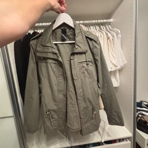 Grön jacka med dragkedja och knappar - Snygg grön jacka med både dragkedja och knappar framtill. Jackan har flera praktiska fickor och axelklaffar som ger en cool look. Perfekt för höst och vår.