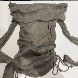 Glittrig off shoulder topp från Gina tricot  - Snygg glittrig off shoulder topp från Gina tricot i en elegant grå nyans. Toppen har en åtsittande passform med knytbara detaljer på sidorna för en justerbar look. Perfekt för en kväll ute eller en speciell tillställning.