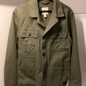 Grön jacka från H&M - Säljer en stilren grön jacka från H&M. Jackan har en klassisk krage och knäppning framtill med knappar. Två bröstfickor med lock och knappar ger en praktisk touch. Perfekt för en avslappnad look.