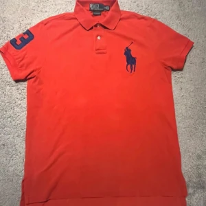 Röd pikétröja från Ralph Lauren - Säljer en snygg röd pikétröja från Ralph Lauren med klassisk krage och knappar. Tröjan har en blå broderad logga på bröstet och en siffra på armen. Perfekt för sommaren som är nu så nära;) (finns ett litet jack på botten)
