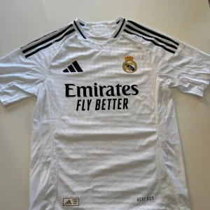 Vit Real Madrid fotbollströja från Adidas - Säljer en vit Real Madrid fotbollströja från Adidas med korta ärmar och svarta detaljer. Tröjan har klubbens emblem på bröstet och sponsorlogga 'Emirates Fly Better'. Perfekt för alla fotbollsfans! ⚽