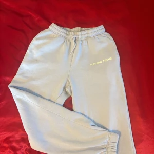 Ljusblåa mjukisbyxor med tryck - Säljer ett par ljusblåa mjukisbyxor med elastisk midja och snörning. Byxorna har ett tryck med texten 'STRNG TGRTHR' på ena benet. Perfekta för en avslappnad stil. 