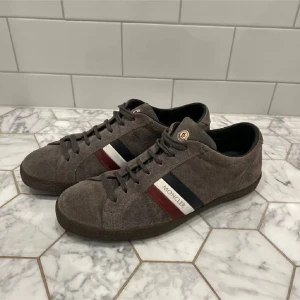 Grå sneakers från Moncler - Snygga grå sneakers från Moncler i mocka med klassiska ränder i rött, vitt och blått på sidan. Skorna har snörning och en rund tå, perfekt för en stilren look.