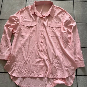 Rosa skjorta med bröstfickor - Säljer en rosa skjorta med knappar och två bröstfickor. Skjortan är tillverkad i en blandning av bomull och polyester, vilket gör den bekväm och lättskött. Perfekt för en avslappnad stil.