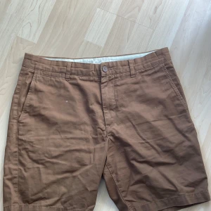 Bruna shorts - Säljer ett par bruna shorts med normal passform. De har en klassisk design med knappar och dragkedja framtill samt fickor både fram och bak. Perfekta för en avslappnad stil.