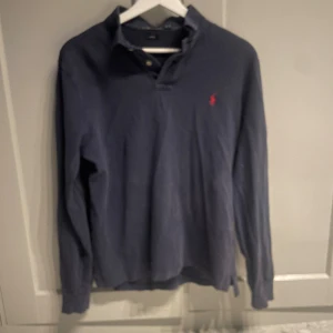 Mörkblå pikétröja från Ralph Lauren - Säljer en stilren mörkblå pikétröja från Ralph Lauren med klassisk krage och knappar. Tröjan har en röd broderad logotyp på bröstet och är i slim fit-modell. Perfekt för en avslappnad men ändå uppklädd look.