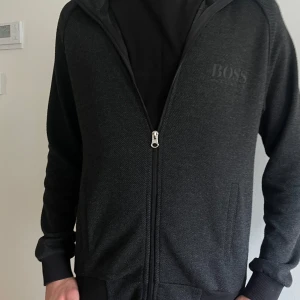 Svart hoodie från Hugo Boss - Snygg svart half zip  från Hugo Boss med dragkedja och ribbade muddar. Perfekt för en stilren look. Diskret logga på bröstet och bekväma fickor framtill.