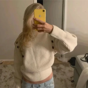 Vit ribbad tröja med knappar - Säljer en stilren vit ribbad tröja med dekorativa knappar på axlarna. Tröjan har en croppad passform och långa ärmar, perfekt för en trendig look. Passar bra till jeans eller kjol.