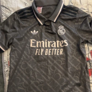 Grå fotbollströja från Adidas - Säljer en grå fotbollströja från Adidas med Real Madrid-logga och 'Emirates Fly Better' tryckt på framsidan. Tröjan har korta ärmar och en klassisk krage med vita detaljer. Perfekt för fotbollsfans!