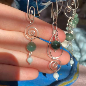4 olika sorters handgjorda örhängen med gröna stenar - Unika handgjorda örhängen med gröna stenar och silverfärgade detaljer. Designen har en elegant spiralform och små pärlor som ger en bohemisk känsla. Perfekt för att lägga till en touch av naturinspirerad stil till din look. 100 kr för ett par. På bilden visas ett örhänge av varje design. 