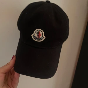 Svart keps från Moncler - Snygg svart keps från Moncler med broderad logga framtill. Justerbar rem med röd, vit och blå detalj baktill. Perfekt för en stilren look.orginal!!!