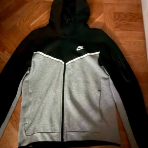 Grå och svart hoodie från Nike - Säljer en snygg grå och svart hoodie från Nike med dragkedja och huva. Perfekt för en sportig look eller avslappnade dagar. Den har långa ärmar och en liten Nike-logga på bröstet.