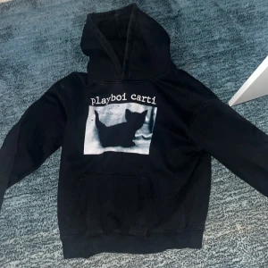 Svart hoodie med tryck - Säljer en svart hoodie med tryck av en siluett och texten 'playboi carti' på framsidan. Tröjan har en klassisk huva och en stor ficka framtill. Perfekt för en avslappnad stil.