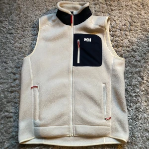 Vit fleeceväst från Helly Hansen - Säljer en vit fleeceväst från Helly Hansen med dragkedja och en praktisk bröstficka i marinblått. Västen har en hög krage och röda detaljer vid dragkedjorna. Perfekt för kyliga dagar! Skick: 9,5/10