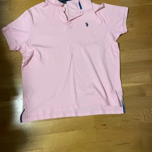 Rosa pikétröja från U.S. Polo Assn. - Säljer en stilren rosa pikétröja från U.S. Polo Assn. med broderad logga på bröstet. Tröjan är kortärmad och tillverkad i 100% bomull för en lyxig känsla. Perfekt för en avslappnad men ändå snygg look.