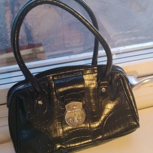 Svart handväska från Complement - Snygg svart handväska i syntetmaterial med krokodilpräglat mönster. Väskan har två handtag och en dekorativ låsdetalj på framsidan. Perfekt för att ge en elegant touch till din outfit. Nästan aldrig andvänd🩷