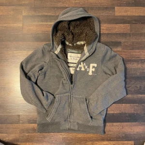 Grå hoodie från Abercrombie & Fitch - Säljer en grå hoodie från Abercrombie & Fitch som är fodrad med päls hela insidan. Jackan har dragkedja och en broderad logga på bröstet. Perfekt för våren samt sommarkvällar.