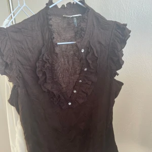 Brun blus från River Island - Säljer en brun blus från River Island med volangdetaljer och knappar framtill. Blusen har korta ärmar och en v-ringad design som ger en elegant touch. Perfekt för att piffa upp garderoben!