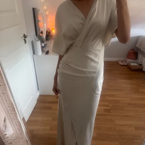 Balklänning - Elegant beige långklänning med omlott-design och v-ringning. Klänningen har kortärmade ärmar och en stilren slits framtill. Helt ny med prislapparna kvar, endast testad. Storlek 34💕💕