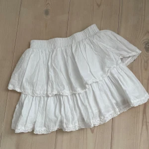 Vit volangkjol - Charmig vit kortkjol med volanger och prickigt mönster. Kjolen har en elastisk midja för bekväm passform och spetsdetaljer längs kanterna. Perfekt för en somrig look!