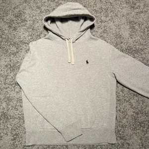 Ralph Lauren hoodie  - Säljer en stilren grå hoodie från Polo Ralph Lauren med en klassisk broderad logga på bröstet. Tröjan har en justerbar huva med snören och en praktisk magficka. Perfekt för en avslappnad look.