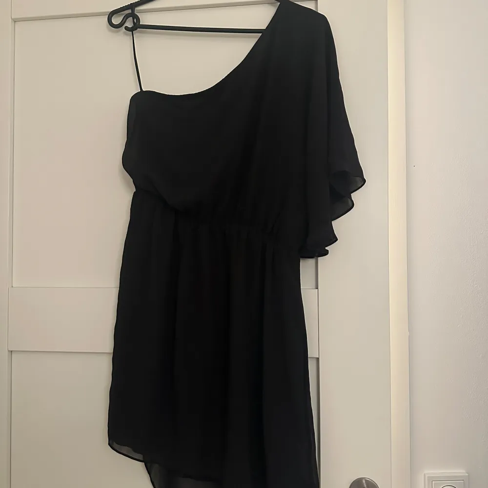 Snygg svart oneshoulder klänning med en luftig design. Perfekt för en kväll ute eller en speciell tillställning. Den har en elegant asymmetrisk stil med en axelbandslös sida och en tunn axelrem på andra sidan.. Mekot.