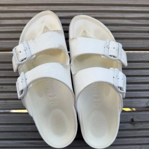 Birkenstock - Säljer ett par vita sandaler från Birkenstock med dubbla justerbara spännen. Perfekta för sommardagar och ger en bekväm passform. Klassisk design som passar till det mesta.