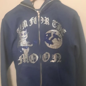 Rhinestone aim for the moon Hoodie - Säljer en blå huvtröja med dragkedja från Aim for the Moon. Tröjan har ett tryck med text och motiv på framsidan. Perfekt för en avslappnad stil.