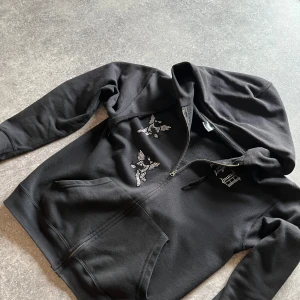 Rhinestone zip - Säljer en svart ziphoodie från Bvaldi med dragkedja och coola detaljer i form av vita tryck på framsidan och text på baksidan. Perfekt för en avslappnad stil. Säljer den pga för liten för mig. 