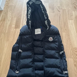 Svart dunväst från Moncler - Snygg svart dunväst från Moncler med huva och dragkedjor. Perfekt för kyligare dagar och ger en stilren look. Moncler-logga på bröstet och märkesdetaljer på huvan.