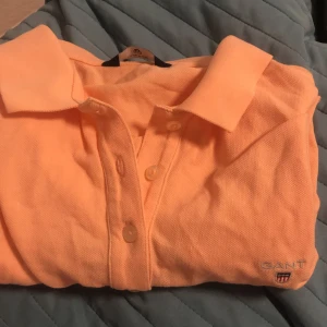 Orange pikétröja från GANT - Säljer en snygg orange pikétröja från GANT. Tröjan har en klassisk krage och knappar framtill. Perfekt för en avslappnad stil med en touch av elegans. den har en liten fläk av penna