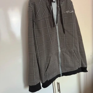  Rip Curl - Säljer en grå rutig hoodie från Rip Curl med dragkedja och huva. Den har ett klassiskt rutmönster och är perfekt för en avslappnad stil. Hoodien har långa ärmar och en bekväm passform.