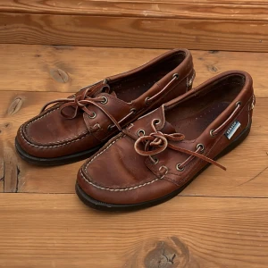 Seglarskor från Sebago - Säljer ett par klassiska bruna seglarskor från Sebago i skinn. Skorna har snörning och en rund tå, perfekt för en stilren look. De har en mörk sula och detaljer i form av metallöljetter.