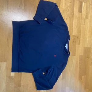 Mörkblå tröja från U.S. Polo Assn. - Säljer en stilren mörkblå tröja från U.S. Polo Assn. med ett diskret rött broderat emblem på bröstet. Tröjan har långa ärmar och en klassisk rund halsringning. Perfekt för en avslappnad och snygg look.