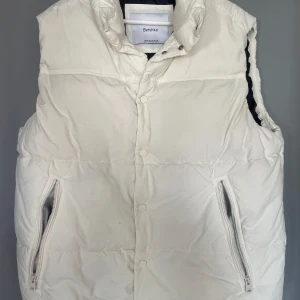 Vit dunväst från Bershka - Säljer en stilren vit dunväst från Bershka. Västen har en hög krage och stängs med tryckknappar. Två praktiska fickor med dragkedja framtill. Perfekt för lager-på-lager under kyligare dagar.
