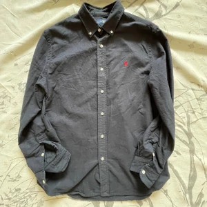 Svart skjorta från Ralph Lauren M - Mörkt Mörkblå skjorta från Ralph Lauren i bomull med klassisk krage och knappar hela vägen. Långärmad modell med ikonisk röd broderad logga på bröstet. Snygg och stilren, passar perfekt till jeans eller chinos.