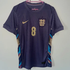 England Alexander-Arnold fotbollströja - Snygg England fotbollströja från Nike med Alexander-Arnold och nummer 8 i guld på ryggen. Tröjan är mörkblå med subtila ränder och har färgglada detaljer i blått, rött och beige längs sidorna. Kortärmad och tillverkad i lätt, ventilerande material för max komfort på planen.