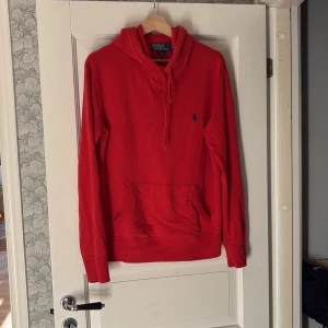 Röd hoodie från Polo Ralph Lauren - Snygg röd hoodie från Polo Ralph Lauren med klassisk blå broderad logga på bröstet. Tröjan har huva med snörning, stor magficka och långa ärmar. Tillverkad i mjuk bomull som är skön att ha på sig. Perfekt för chill dagar eller när du vill ha en clean look. OBS Priset kan diskuteras vid snabb affär!