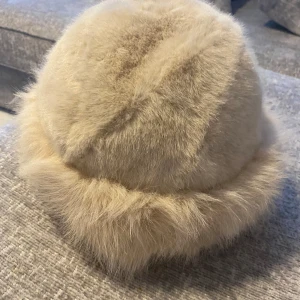 Beige fluffig pälsmössa - Säljer en supermjuk och fluffig beige pälsmössa med rundad form och bred kant. Mössan är helfodrad och har en lyxig vintagekänsla, perfekt för kalla dagar när du vill sticka ut med en cool look. Äkta päls. Aldrig använd. 