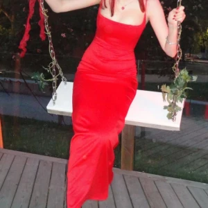 Röd korsett långklänning med smala axelband - Säljer en röd långklänning med smala axelband och figurnära passform. Klänningen har en enkel, stilren design med rak överdel och lätt utsvängd nederdel. Materialet har en lätt glansig finish som ger en lyxig känsla. Perfekt för bal eller festliga tillfällen.