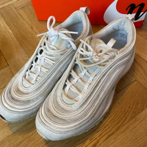 Nike Air Max 97 vita sneakers (storlek 43) - Nike Air Max 97 sneakers i helvitt med ikoniska vågiga linjer och synlig Air-sula. Skorna har mesh och syntetmaterial, rund tå och snörning. Snyggt broderad Swoosh på sidan och klassisk design som sticker ut. Perfekt för dig som gillar streetwear och sportig stil.