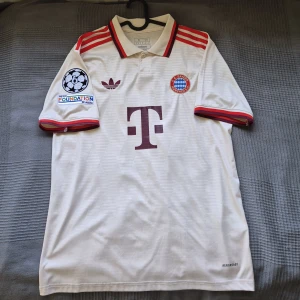 Bayern München 24/25 UCL tröja - Bayern München 24/25 UCL tröja med Musiala 42 på ryggen. Riktigt snygg och klassisk design med retro vibbar. Perfekt att ta till pitchen eller gymmet.