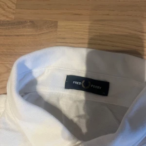 Vit Fred Perry t-shirt med krage XL - Vit t-shirt från Fred Perry i storlek XL med klassisk krage och knappslå framtill. T-shirten har korta ärmar och en bröstficka med Fred Perrys ikoniska lagerkrans-brodyr. Materialet är mjuk bomull som känns skönt mot huden. 
