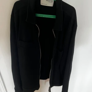 Svart overshirt från Selected Homme - Svart overshirt med klassisk krage och två stora fickor framtill. Jackan har dragkedja och är långärmad, perfekt för lager-på-lager. Materialet är bomull och designen är stilren och enkel, passar dig som gillar minimalistisk look.