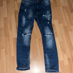 Blå slitna jeans med pärldetaljer - Säljer ett par blå jeans med coola slitningar och hål på knäna, samt små pärldetaljer på framsidan och ena benet. Jeansen har klassisk femficksdesign och en rak passform som ger en avslappnad vibe. Perfekt för dig som gillar streetstyle och vill sticka ut.