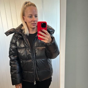Svart pufferjacka med huva - Säljer en svart pufferjacka med glansig finish och stor huva. Jackan har breda, quiltade sektioner och tvåvägsdragkedja framtill. Passformen är loose och jackan är perfekt för kalla dagar. Snygg och trendig look som håller dig varm hela vintern. NY aldrig använd endast privat, tagg kvar! 
