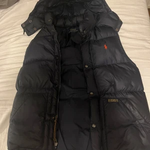 Mörkblå dunväst från Polo Ralph Lauren - Mörkblå dunväst från Ralph Lauren med klassisk röd logga på bröstet. Västen har hög krage, dragkedja och tryckknappar framtill samt två stora fickor. Perfekt för lager-på-lager och riktigt snygg till hoodie eller stickad tröja.