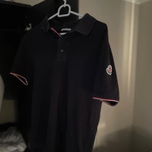 Svart Moncler piké - Svart piké från Moncler med klassisk polokrage och tre knappar framtill. På vänster ärm sitter Monclers ikoniska logga. Ärmarna har snygga rödvita detaljer längs kanten.                                Pris kan såklart diskuteras✅