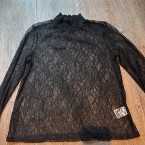 Svart spetsig polotopp från Levete - Supersnygg svart polotopp från Levete i storlek S. Hela toppen är gjord i transparent spets med ett coolt mönster och har långärmade ärmar. Perfekt att styla lager på lager eller med ett linne under för extra edge.